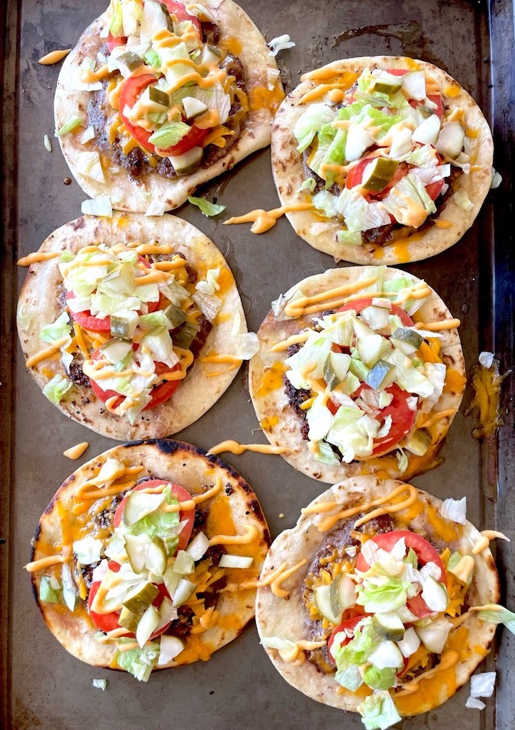 Six Smashed Cheeseburger Tacos on a sheet pan loaded with burger condiments such as lettuce, tomato, pickle, and Bic Mac Sauce. 