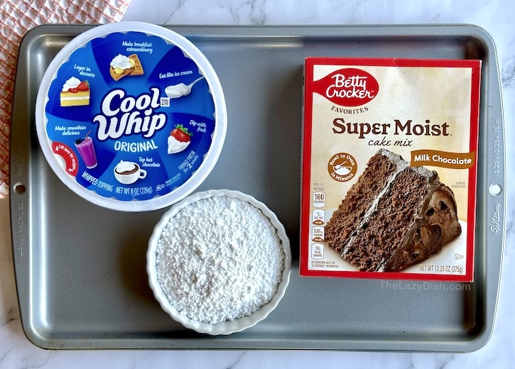 Sheet pan topped with ingredients to make Cool Whip Cookies: a box of Betty Crocker chocolate cake mix, powdered sugar, and a tub of Cool Whip.