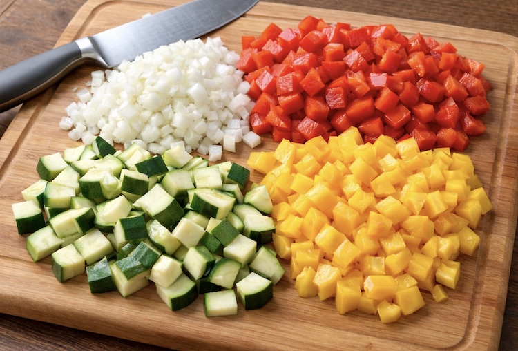 A wooden cutting board full of freshly chopped onion, colorful bell peppers, and zucchini. 