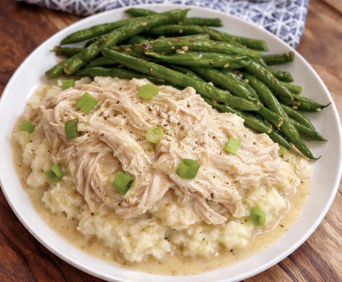 Close up view of shredded chicken and gravy over mashed potatoes with a side of sautéed green beans. This easy dinner is made in a crockpot with common ingredients for simple weeknight meals. 