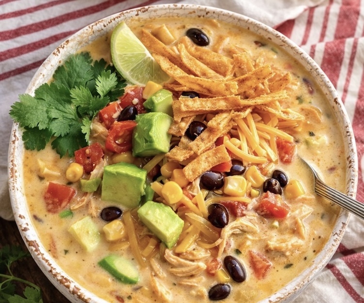 Close up view of creamy chicken taco soup in a bowl garnished with tortilla chips, avocado, lime, and cilantro. 